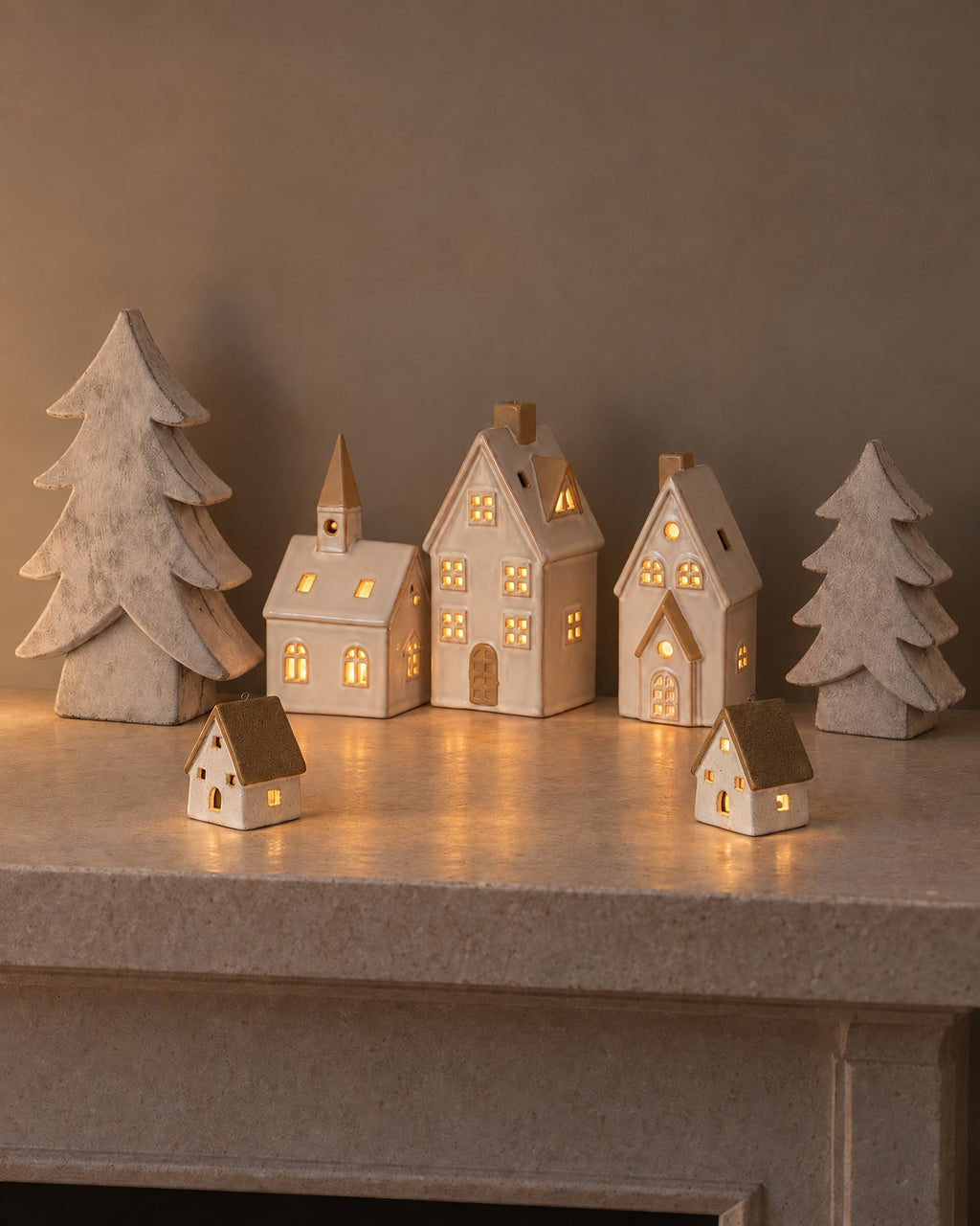 The Little Christmas Village on a stone mantel: the Alora ceramic church, two townhouses and two cottages glowing with LED lights between two stone-effect Athena Christmas trees
