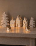 The Little Christmas Village on a stone mantel: the Alora ceramic church, two townhouses and two cottages glowing with LED lights between two stone-effect Athena Christmas trees