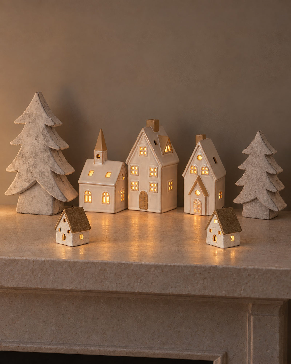 The Little Christmas Village on a stone mantel: the Alora ceramic church, two townhouses and two cottages glowing with LED lights between two stone-effect Athena Christmas trees