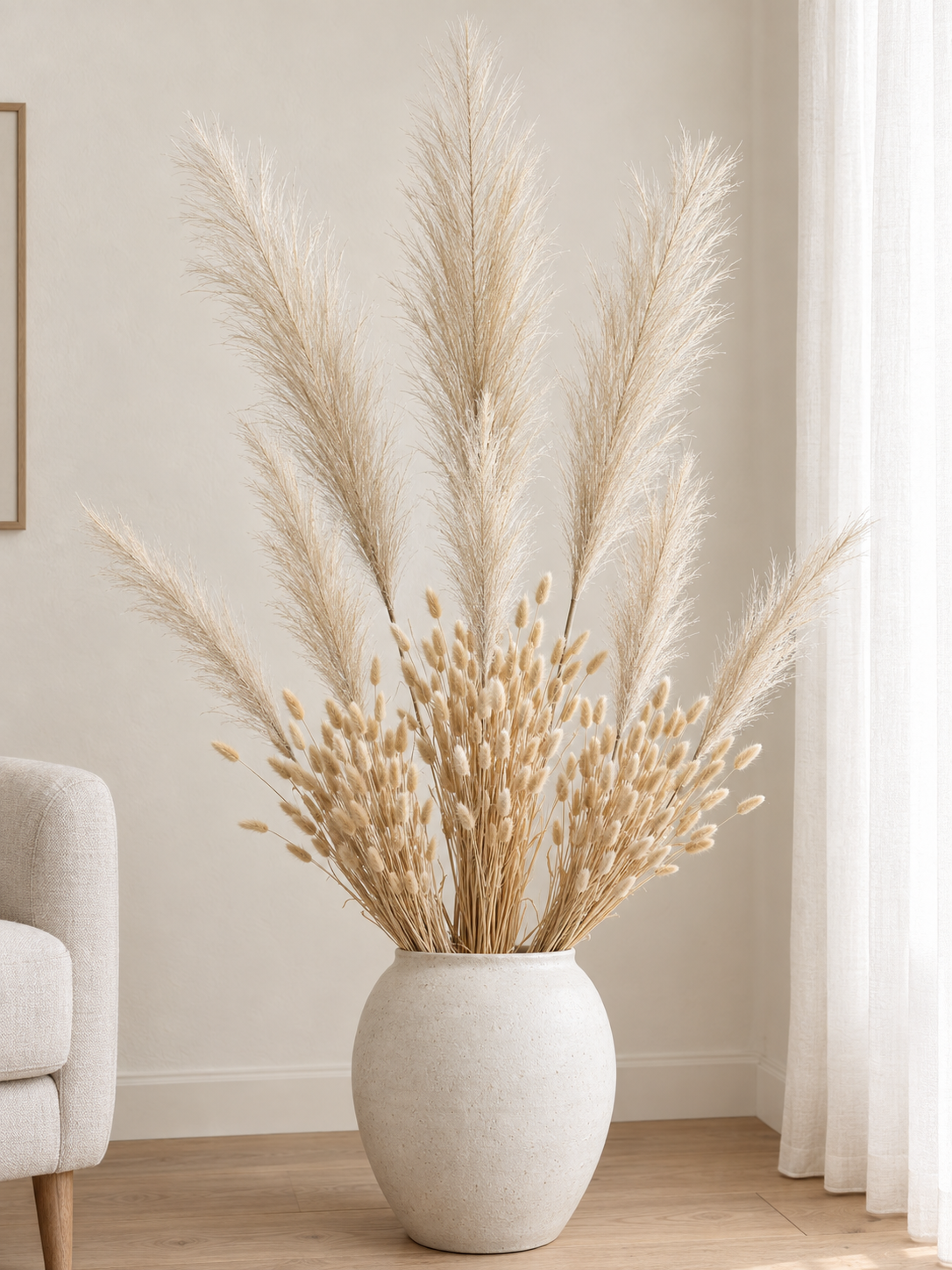 The Bleached Meadow Bouquet with large and smaller bleached faux pampas plumes and natural dried bunny tails in a chalk-white styling vase beside a linen chair; vase not included