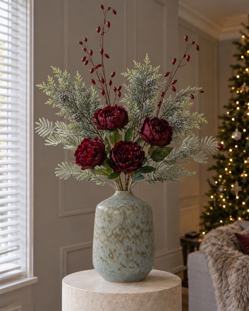 The Capella Burgundy Christmas Arrangement: artificial burgundy peony roses, burgundy rosehips, silver wattle and frosted pine in the blue-grey Capella Bottle Vase, on a stone plinth in a Christmas living room