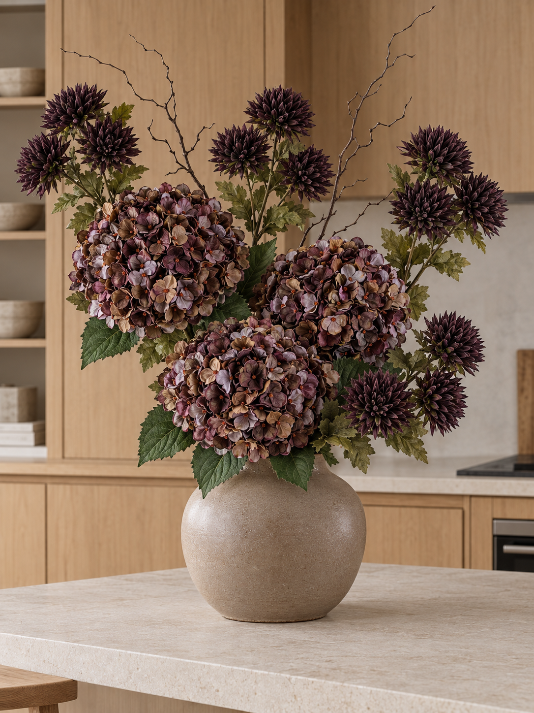 The Cocoa Autumn Bouquet with variegated plum and coffee hydrangeas, dark chocolate chrysanthemums and bare branches, styled in a taupe vase on a kitchen island; vase not included