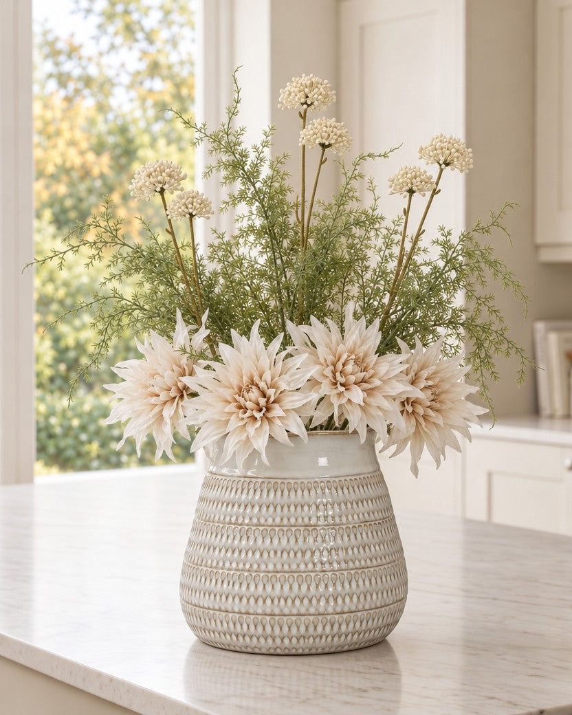 The Cotswold Cream Bouquet: artificial Café au Lait dahlias resting at the rim of the wide Tava Wide Hewn Vase from the With vase option, with cream allium sprays and asparagus fern rising above, on a marble kitchen island
