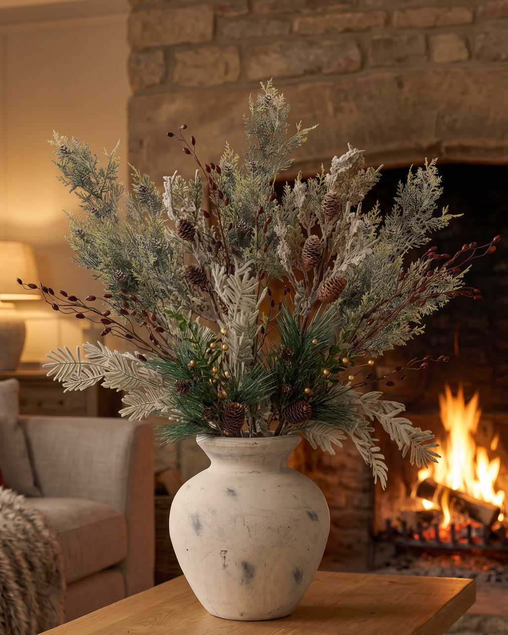 The artificial Darcy Antique White Christmas Arrangement on an oak coffee table in front of a lit stone fireplace