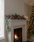 The Frosted Pine Mantel Garland Set draped along a pale grey mantel above a lit fire, with the artificial red berry and gold sprays tucked in along the garland