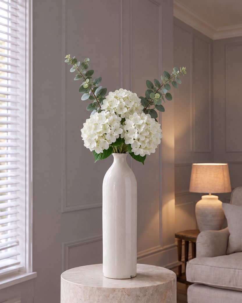 The Gisela White Arrangement: three artificial white hydrangeas and two eucalyptus sprays in the tall cream crackle-glazed Garda Gisela bottle vase, on a stone plinth in a panelled living room