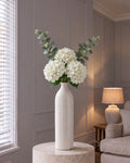The Gisela White Arrangement: three artificial white hydrangeas and two eucalyptus sprays in the tall cream crackle-glazed Garda Gisela bottle vase, on a stone plinth in a panelled living room