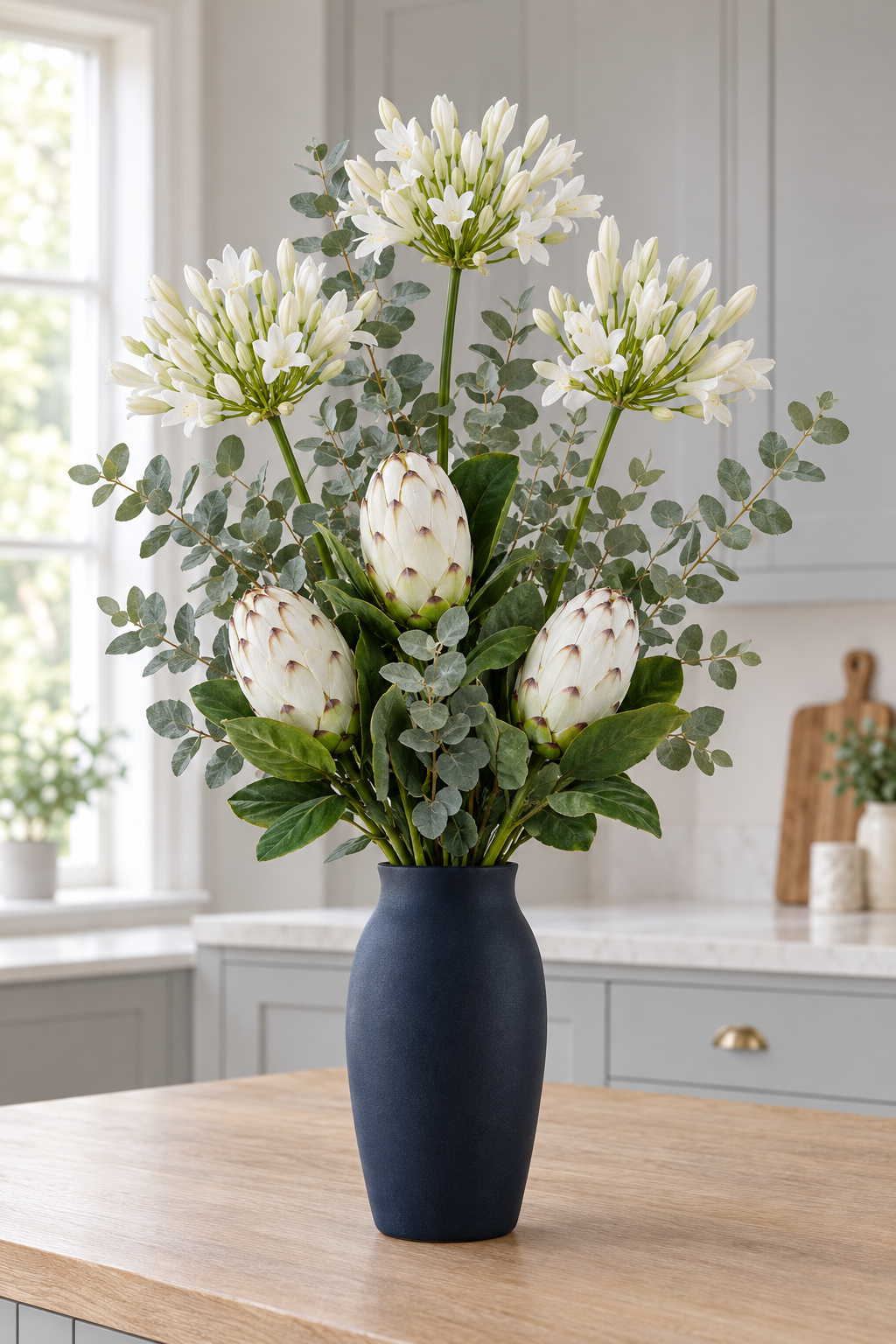 The Ivory Protea Bouquet with three closed white protea heads, three cream agapanthus sprays and cineraria eucalyptus in a dark navy styling vase on a kitchen island; vase not included