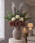 The Mocha and Cream Hydrangea Bouquet in a ribbed brown amphora vase with handles on a stone plinth, with artificial coffee and cream hydrangeas, grey pampas and seeded eucalyptus, in a panelled living room