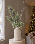 The Monte Winter White Arrangement: artificial frosted pine, a snowy woodland branch and a lambs ear and waxflower sprig in the white ribbed Monte Large Bell Vase, on a stone plinth in a panelled living room beside a lit Christmas tree