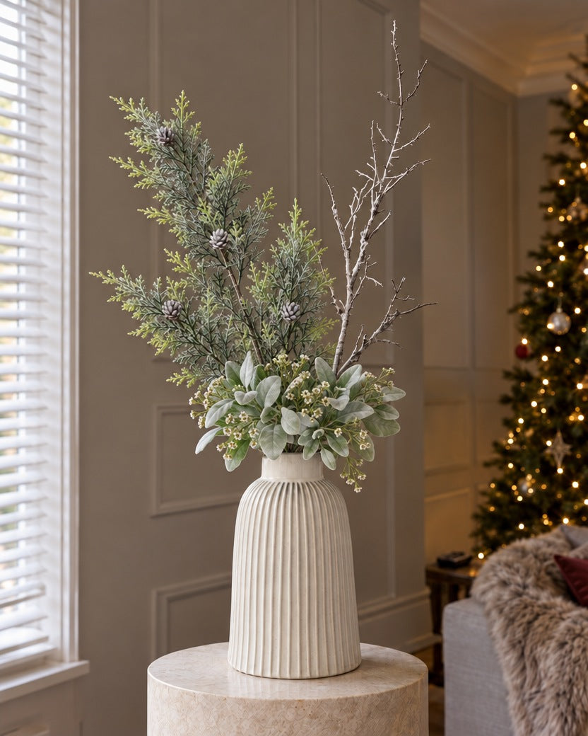 The Monte Winter White Arrangement: artificial frosted pine, a snowy woodland branch and a lambs ear and waxflower sprig in the white ribbed Monte Large Bell Vase, on a stone plinth in a panelled living room beside a lit Christmas tree