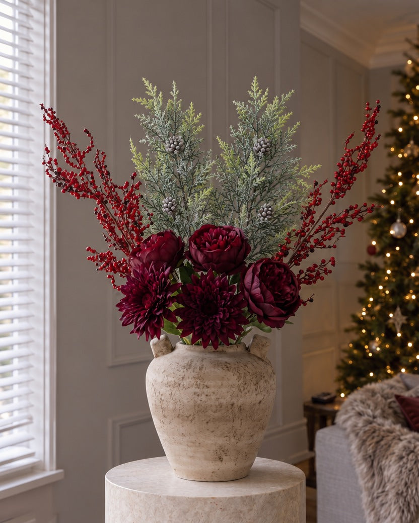 The Mulled Burgundy Christmas Bouquet in the Terra urn vase from the With vase option: artificial burgundy peony roses, dark dahlias, red berry sprays and frosted pine, on a stone plinth beside a lit Christmas tree