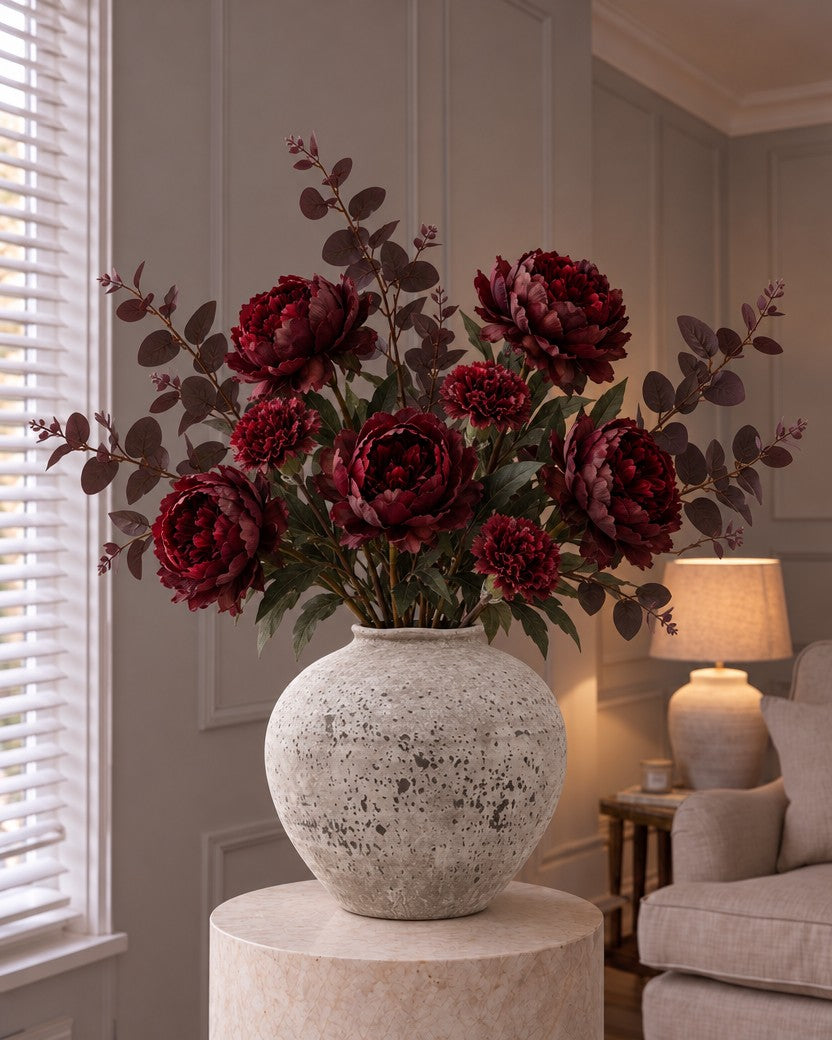 The Nocturne Burgundy Bouquet: five artificial burgundy peony roses, frilled burgundy carnations and wine-toned eucalyptus in the aged grey Regola Stone Ceramic Vase from the With vase option, on a stone plinth in a lamplit panelled living room