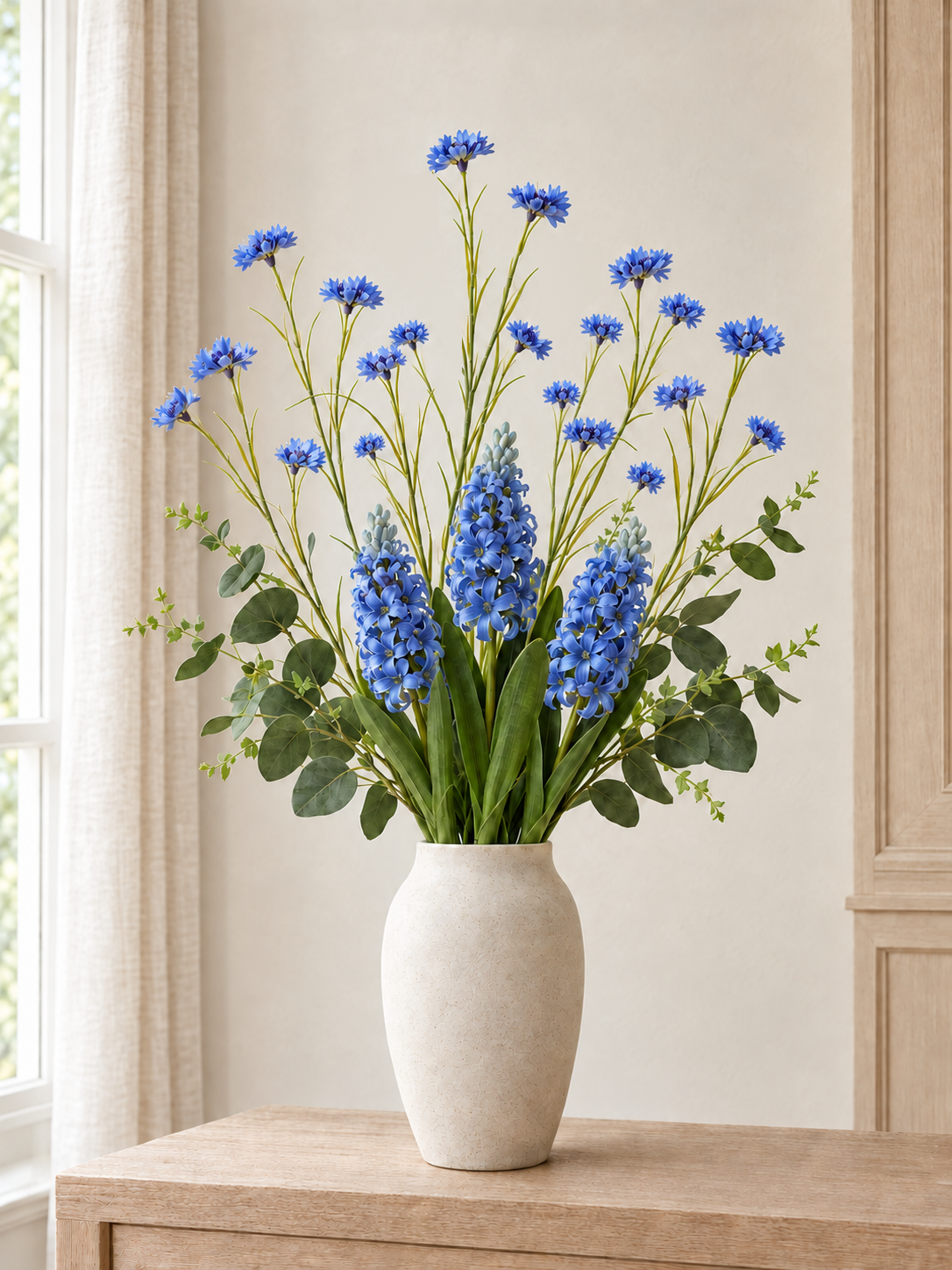 The Provence Meadow Bouquet with branching blue cornflowers, three blue hyacinths and green eucalyptus in a neutral styling vase beside a window; vase not included