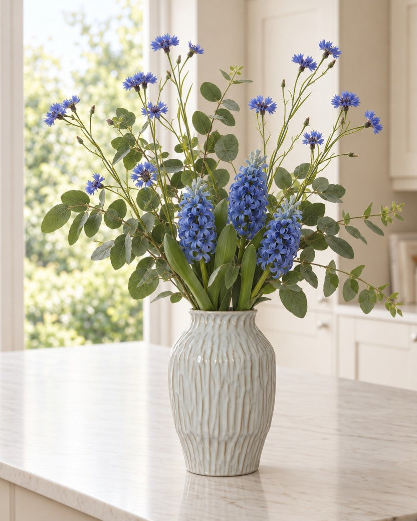The Provence Meadow Bouquet: artificial blue cornflowers, three blue hyacinths and green eucalyptus in the rippled white Numa Olpe Vase from the With vase option, on a marble kitchen island