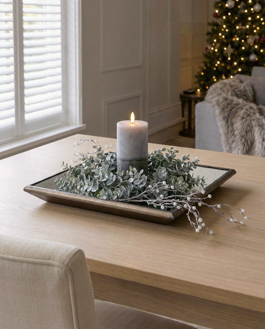 The Lit Frosted Eucalyptus Hearth Tablepiece on an oak dining table in a panelled living room: mirrored tray, artificial frosted eucalyptus wreath, stone LED candle and pearl spray