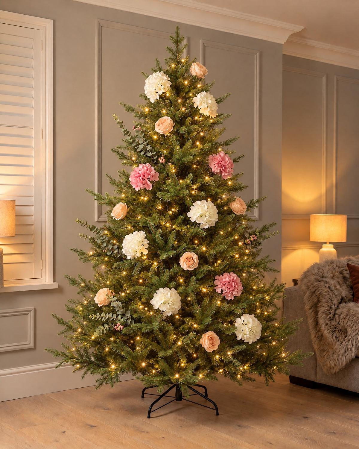 The Winter Bloom Flower Christmas Tree, 7ft Spruce (210cm tall, 140cm wide), lit in a panelled living room in the evening, dressed with artificial white and pink hydrangeas, blush roses and eucalyptus