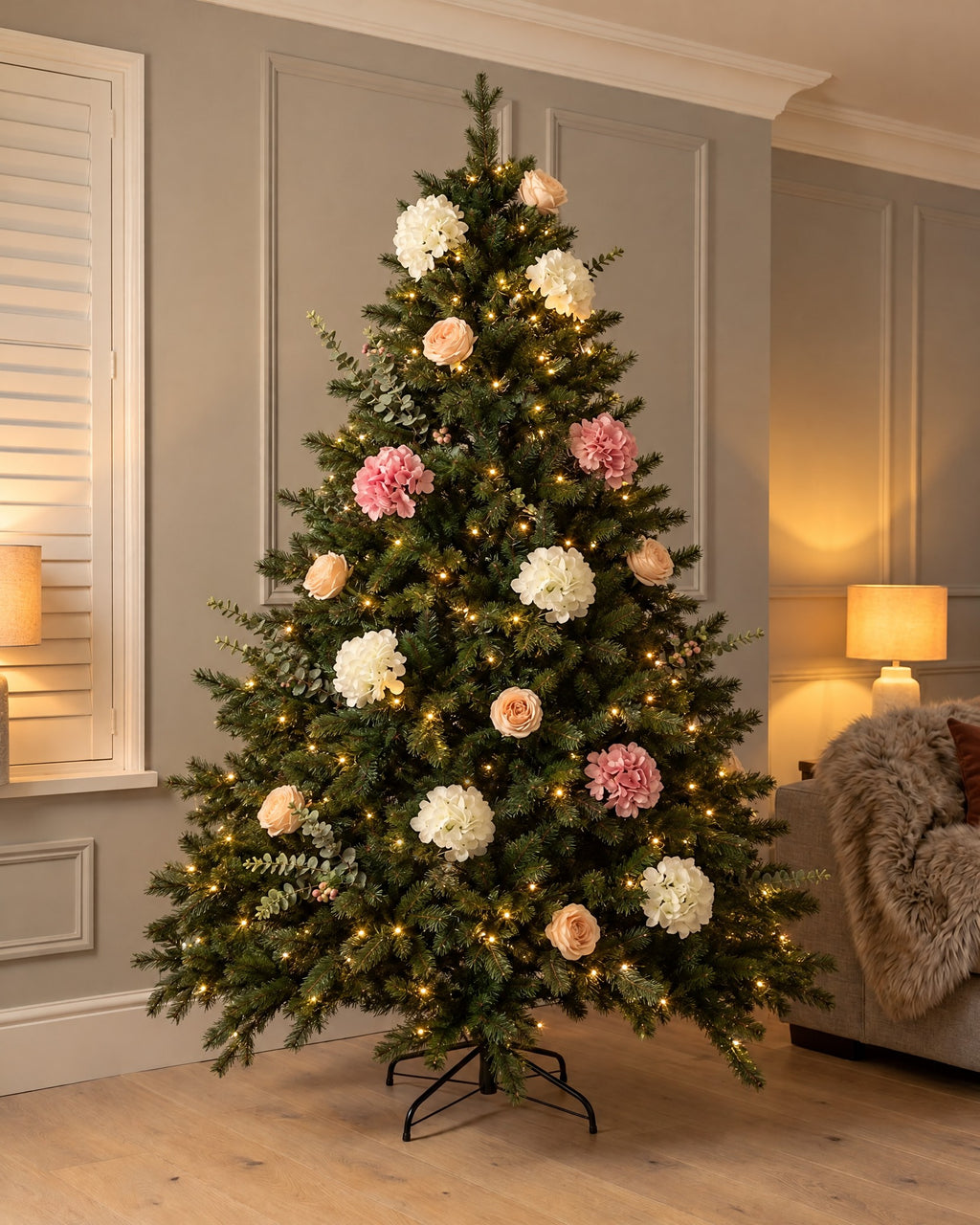 The Winter Bloom Flower Christmas Tree, 7ft Forest Fir (210cm tall, 140cm wide), lit in a panelled living room in the evening, dressed with artificial white and pink hydrangeas, blush roses and eucalyptus