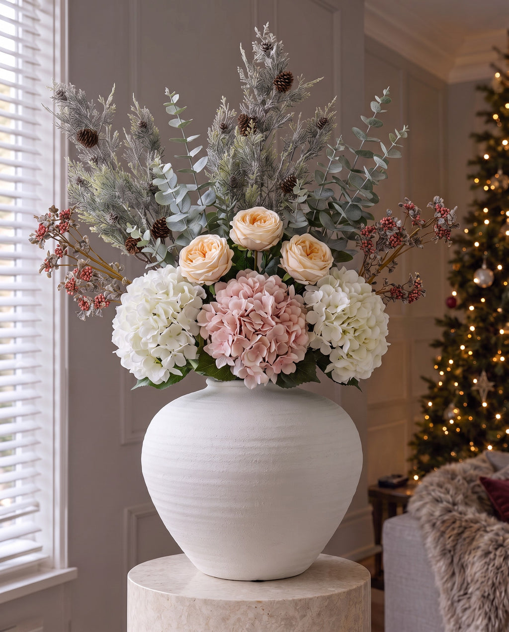 The Winter Bloom Bouquet in the ribbed matt white Regola vase on a stone plinth, with artificial blush roses, white and pink hydrangeas, pink berries, frosted pine and eucalyptus, in a living room with a lit Christmas tree