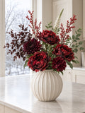The Winter Claret Bouquet in the Grove Globe Vase from the With vase option: artificial burgundy peony roses, a raven dahlia, red winter berry branches, burgundy and green eucalyptus and a white veronica spike, on a marble kitchen island by a winter window