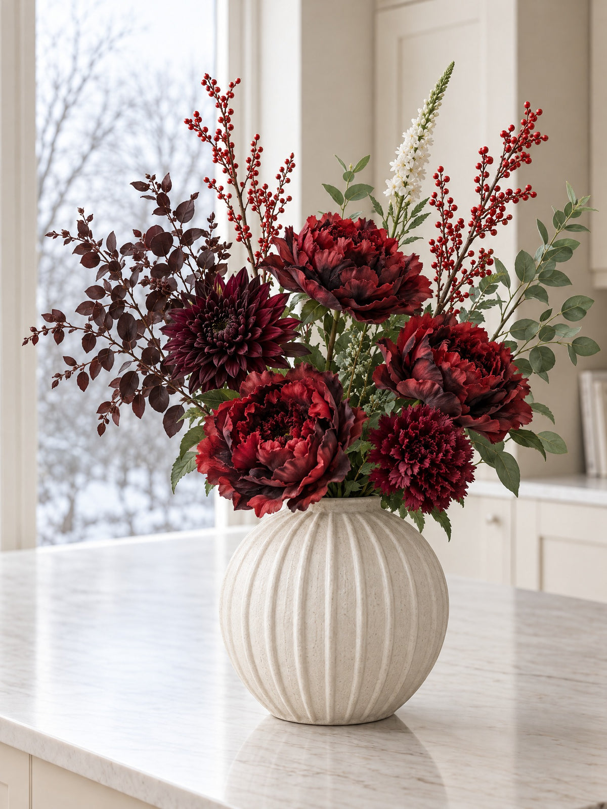 The Winter Claret Bouquet in the Grove Globe Vase from the With vase option: artificial burgundy peony roses, a raven dahlia, red winter berry branches, burgundy and green eucalyptus and a white veronica spike, on a marble kitchen island by a winter window