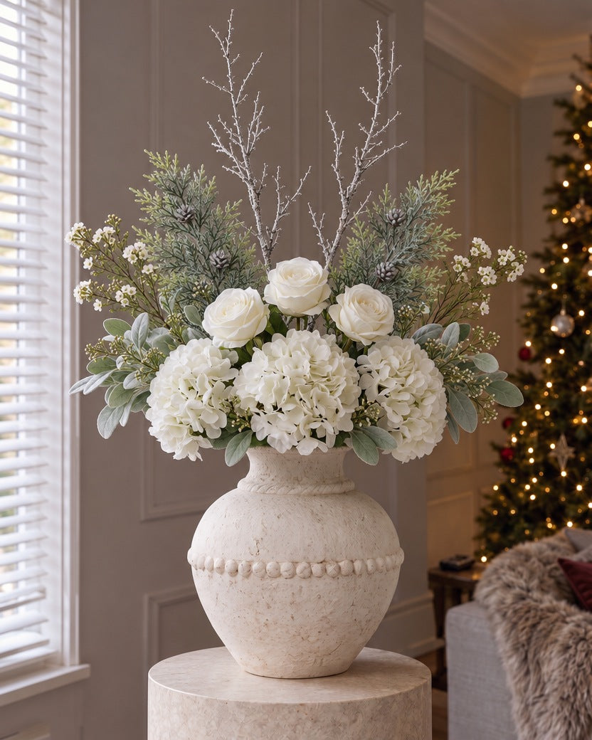 The Winter White Christmas Bouquet: artificial white hydrangeas, white roses, frosted pine, snowy branches and waxflower in the Large Olympia vase from the With vase option, on a stone plinth in a panelled living room beside a lit Christmas tree