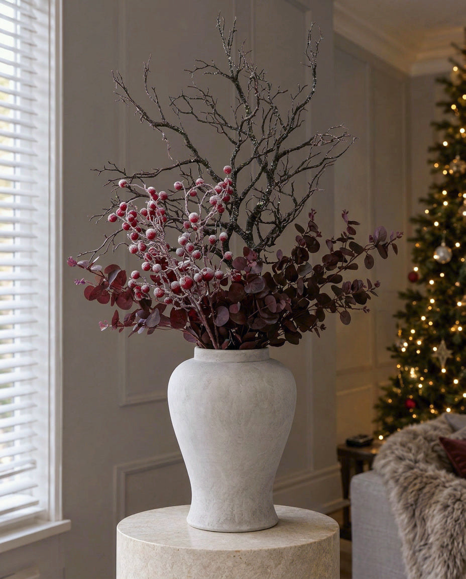 The Winter Woodland & Frosted Berry Urn in the Darcy stone ginger jar on a stone plinth in a panelled living room, with an artificial speckled branch, frosted red berries and burgundy eucalyptus
